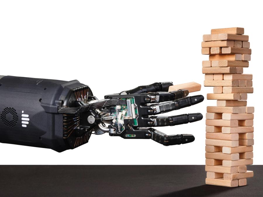 A black prosthetic hand that imitates human fingers and tendons holds a wooden block near a tower of blocks.
