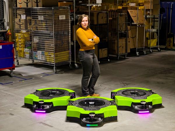 A blonde woman in a yellow shirt and black jeans stands in a warehouse. Three wide, low to the ground green and black Amazon warehouse robots are in front of her.