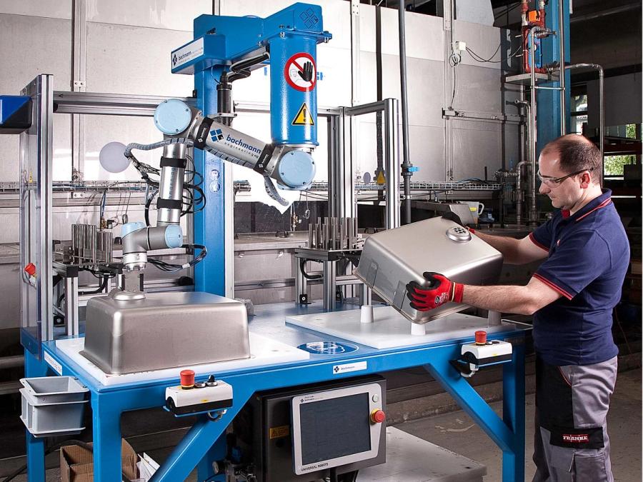 The grey and blue arm is seen attached to an overhead joint in tabletop setup in a factory. The robot manipulates a sink, while a worker examines another sink.