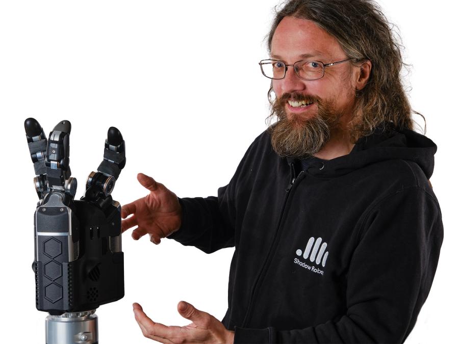 A man with long hair, a beard and mustache, and glasses smiles while gesturing towards the DEX-EEE robotic hand.