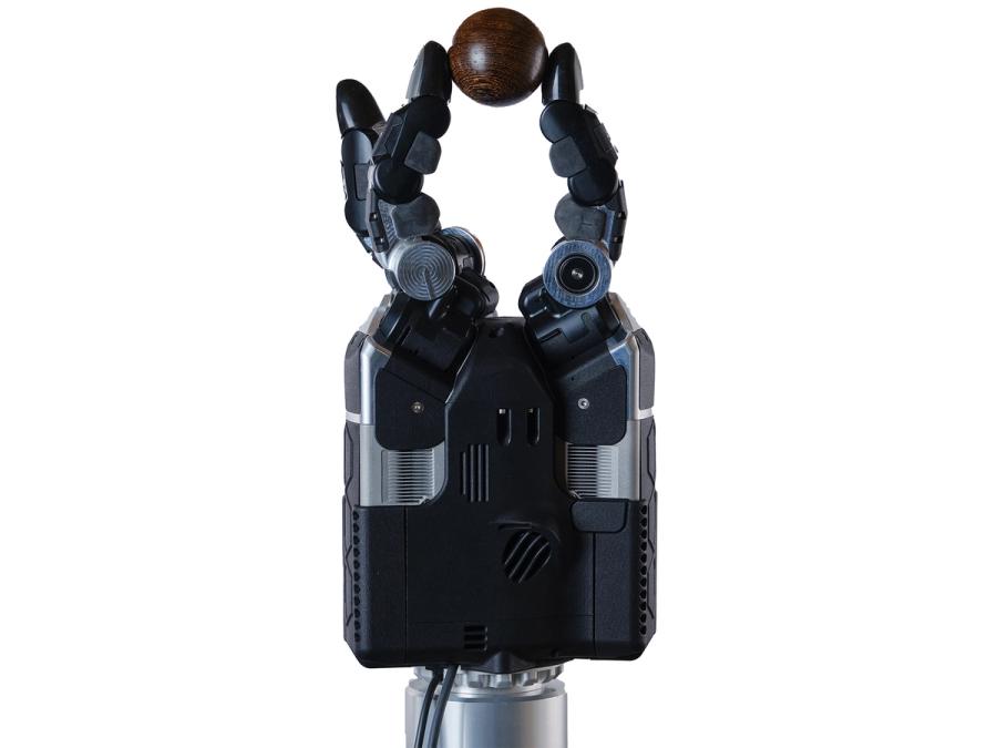 The three fingered black and silver robotic gripper hand DEX-EE pinches a small brown ball between two fingers.