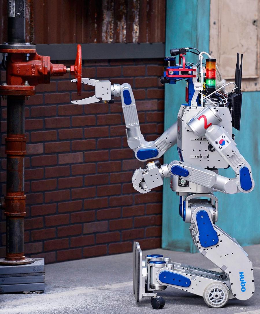 A bipedal robots legs are folded forward so that it can kneel, while it's gripper hand spins a hand wheel attached to a pole.