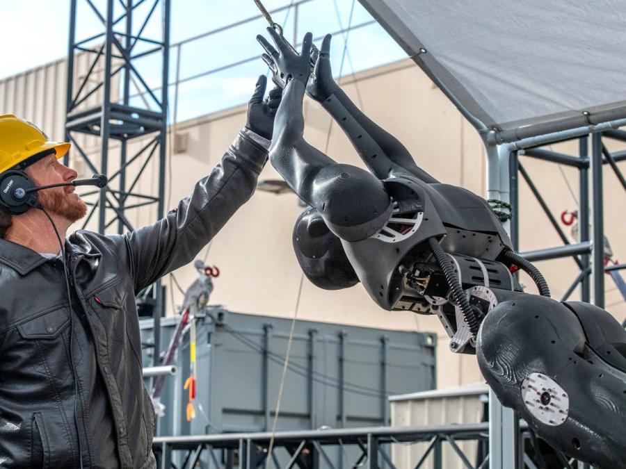 A man in a yellow hardhat and headset prepares a grey humanoid, which is bent over with its hands raised over its head.