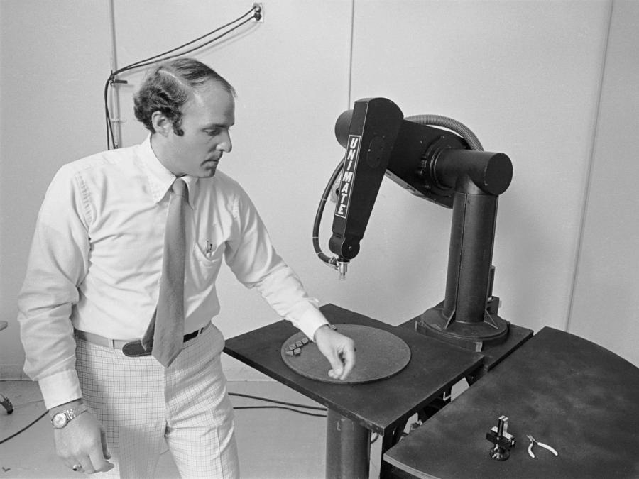 A black and white photo of a business man in front of a tabletop black industrial robot arm.
