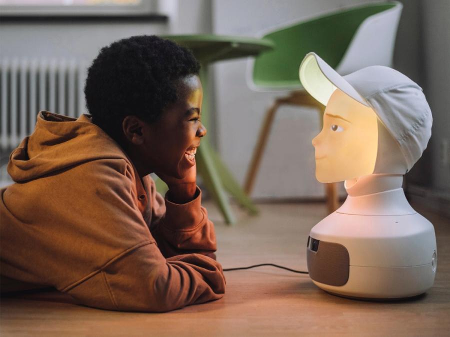 A smiling boy lies on the floor interacting with a Furhat social robot, which has a glowing backlit projected face.