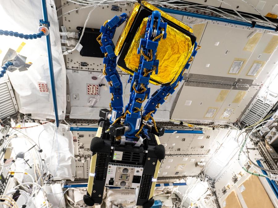 Astrobee, a toaster-sized floating space robot with 4 blue grippers holding a golden cube, while it flies on the International Space Station.