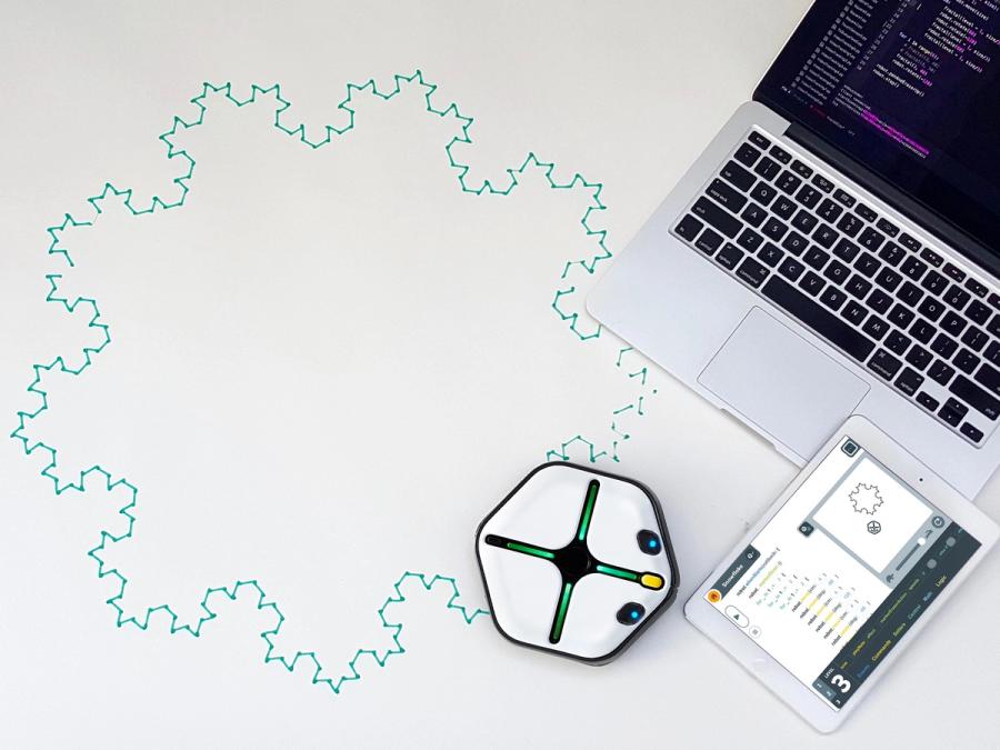 A squat white hexagonal robot with glowing lights sits in front of a tablet and laptop. It has drawn a large shape like a snowflake.