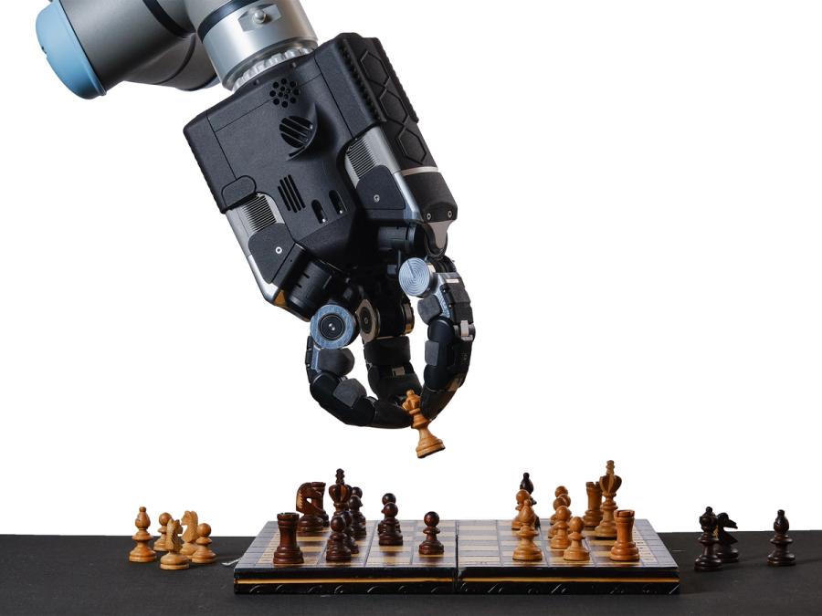 A three fingered black and silver robotic gripper hand manipulates a chess piece onto a board.