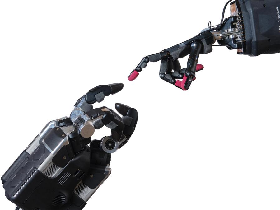 A three fingered robotic gripper hand reaches a finger out to a five fingered robotic hand.