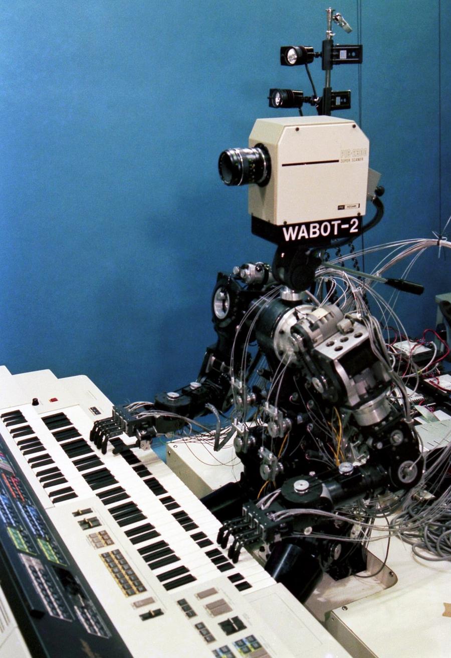 A historical robot with a complex humanoid body and a 1980's era video camera for a head sits and plays the keyboards.
