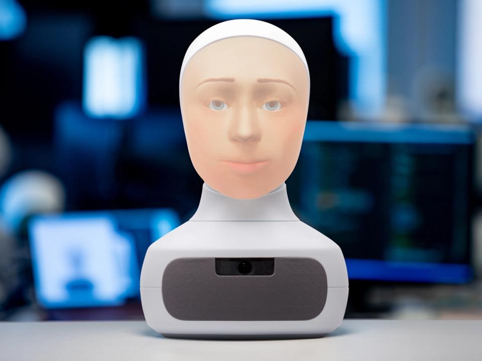 A Furhat robot head sits on a desk. It has a white base and a back projected face.