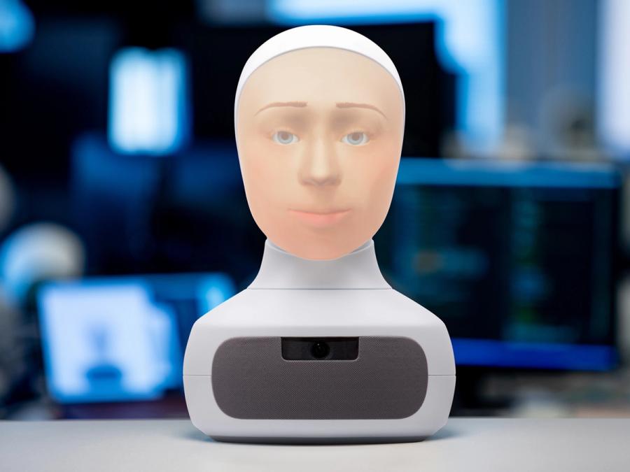 A Furhat robot head sits on a desk. It has a white base and a back projected face.