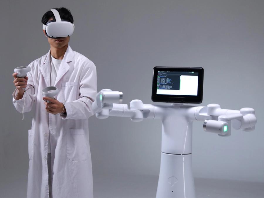 A person in a lab coat and VR equipment stands next to a wheeled humanoid robot with a tablet display for a head.