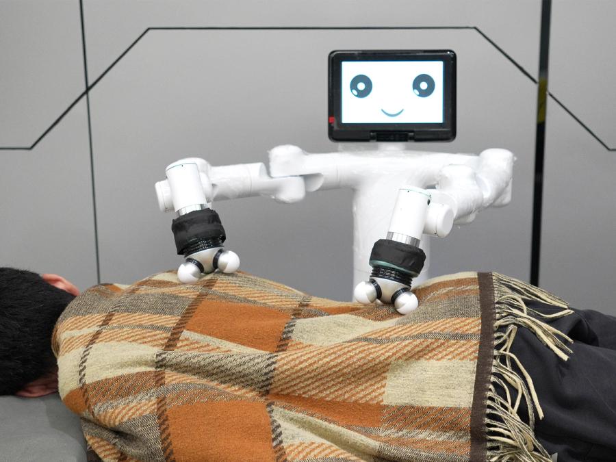 Mercury X1, a wheeled humanoid robot with a tablet display head, uses its two robotic arms with attachments for massaging on a person lying on a table.