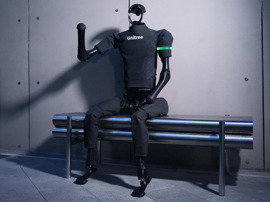 A gray bipedal humanoid wearing a Unitree shirt sits on a bench, with one arm raised.