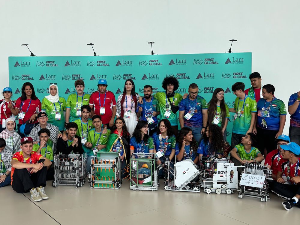 A group of students and adults from different countries pose with robots in front of signage for the First Global robotics competition.